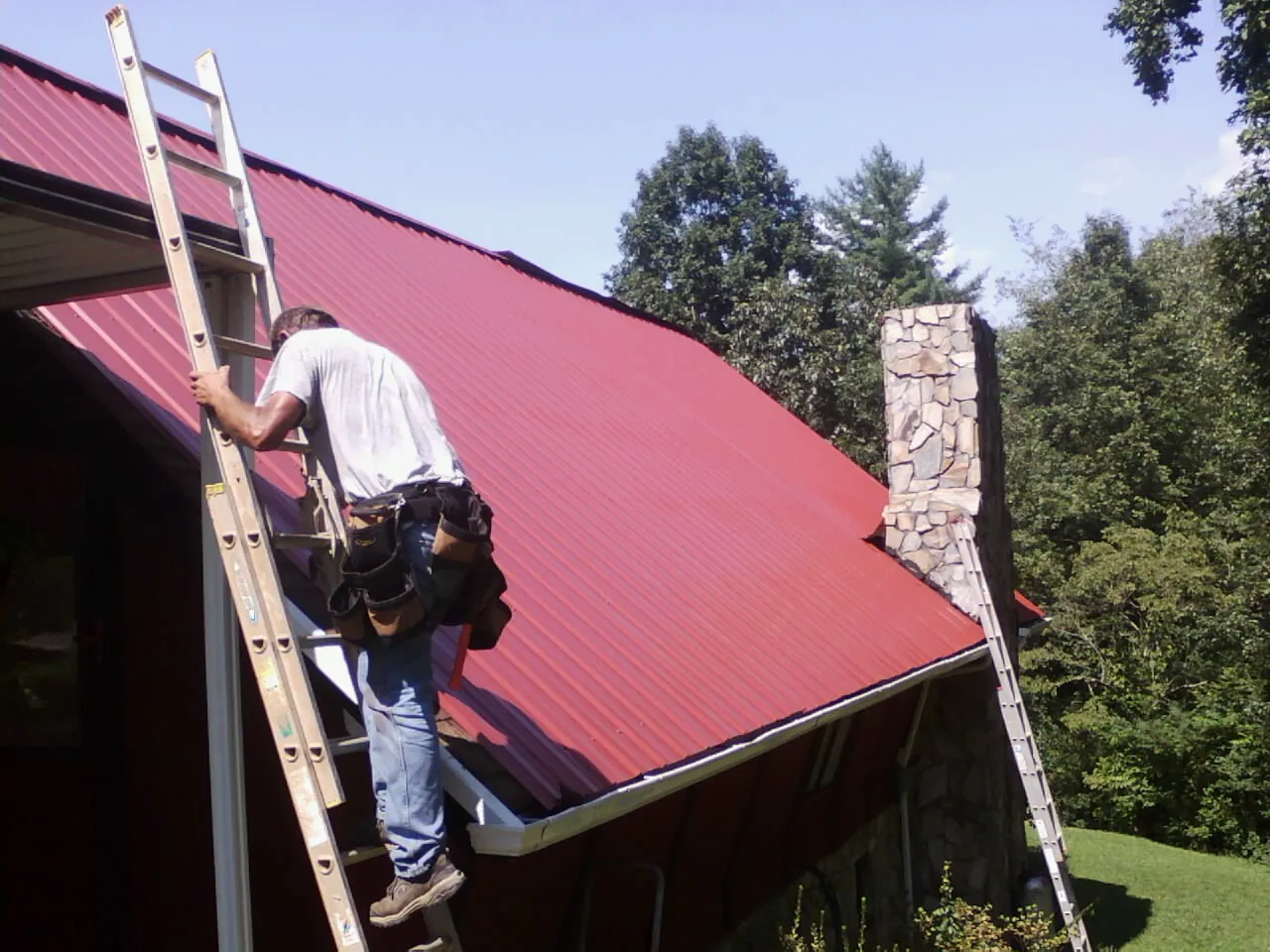 Roofer on a ladder beside a red metal roof and stone chimney — Soffit & Fascia Repair by Roofing Brothers in Beacon Falls